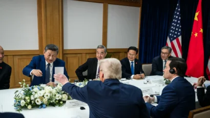 Leaders from the United States and China sit around a conference table with documents, flowers, and flags, engaging in a formal discussion.