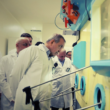 A group of men in white lab coats observe equipment inside a laboratory.