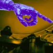 A large blue robotic hand points towards a person seated at a desk in a grand, ornate room with yellow lighting.