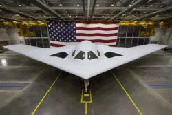 A sleek, white stealth bomber aircraft is displayed in a hangar with a large American flag in the background.