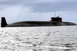 A large, black military submarine is partially submerged and moving through calm waters under a cloudy sky.