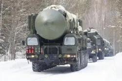 Military trucks carrying large missile systems drive on a snow-covered road through a forested area.