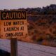 Sign reading "Caution Low Water Launch At Own Risk" at Great Salt Lake marina during a drought in front of naturally-colored grasses that look like fire.